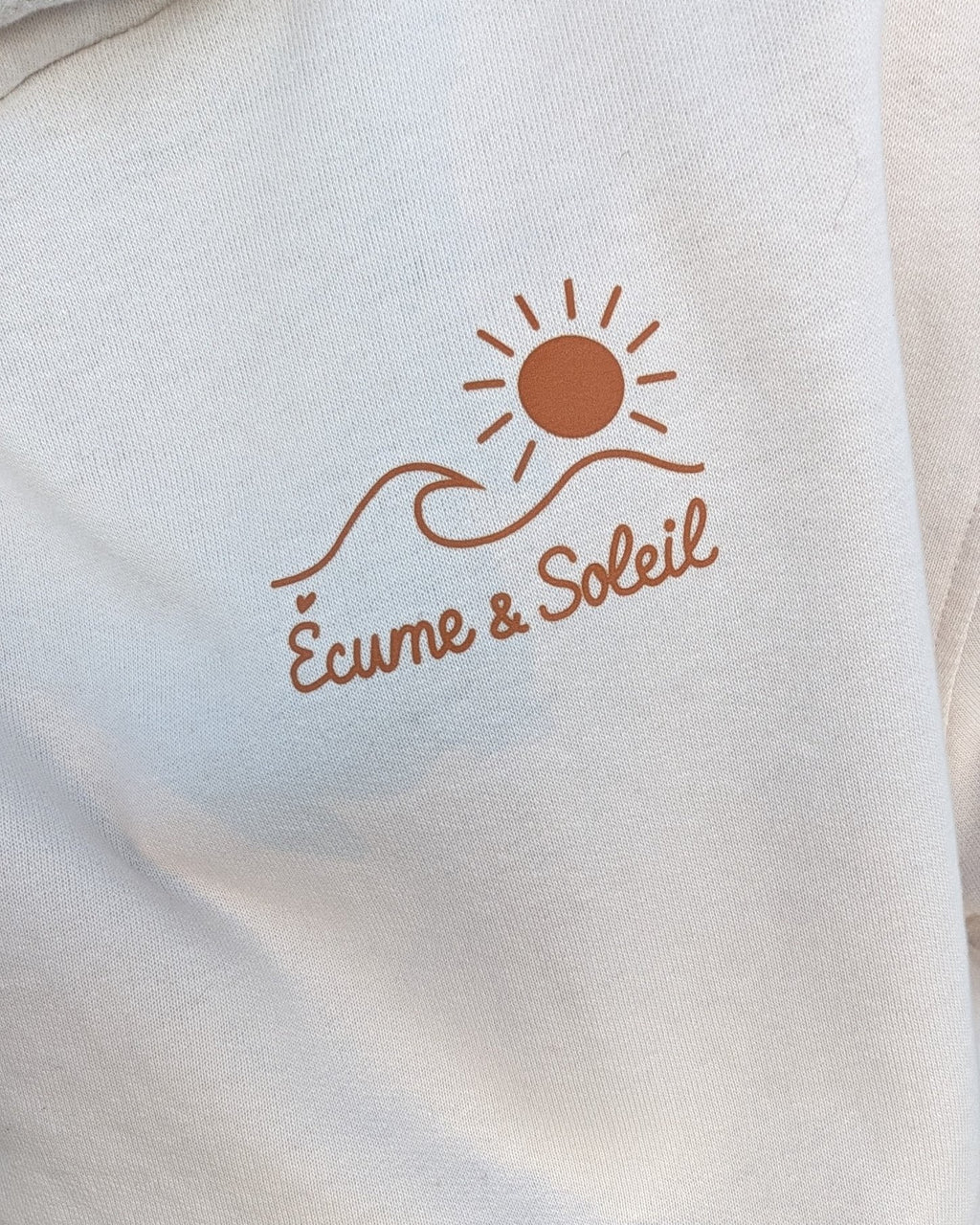 Gros plan du logo Écume & Soleil marqué en terracotta au cœur du sweat à capuche écru