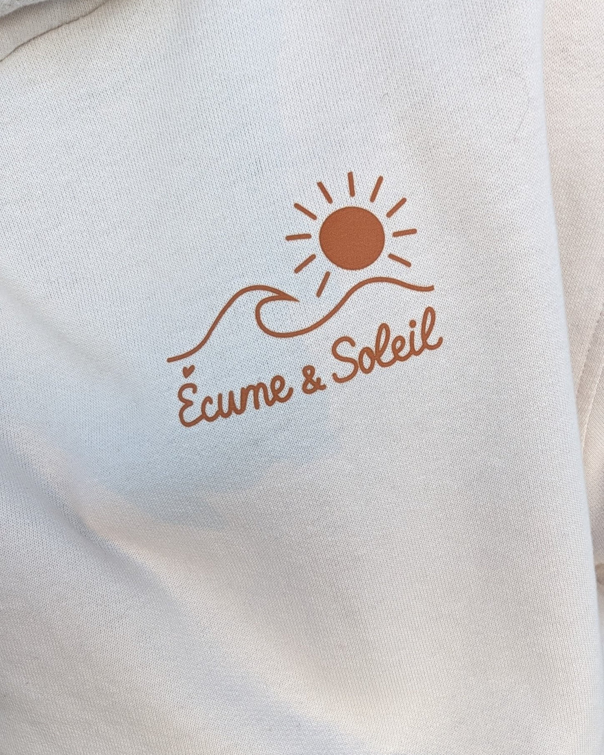 Gros plan du logo Écume & Soleil marqué en terracotta au cœur du sweat à capuche écru