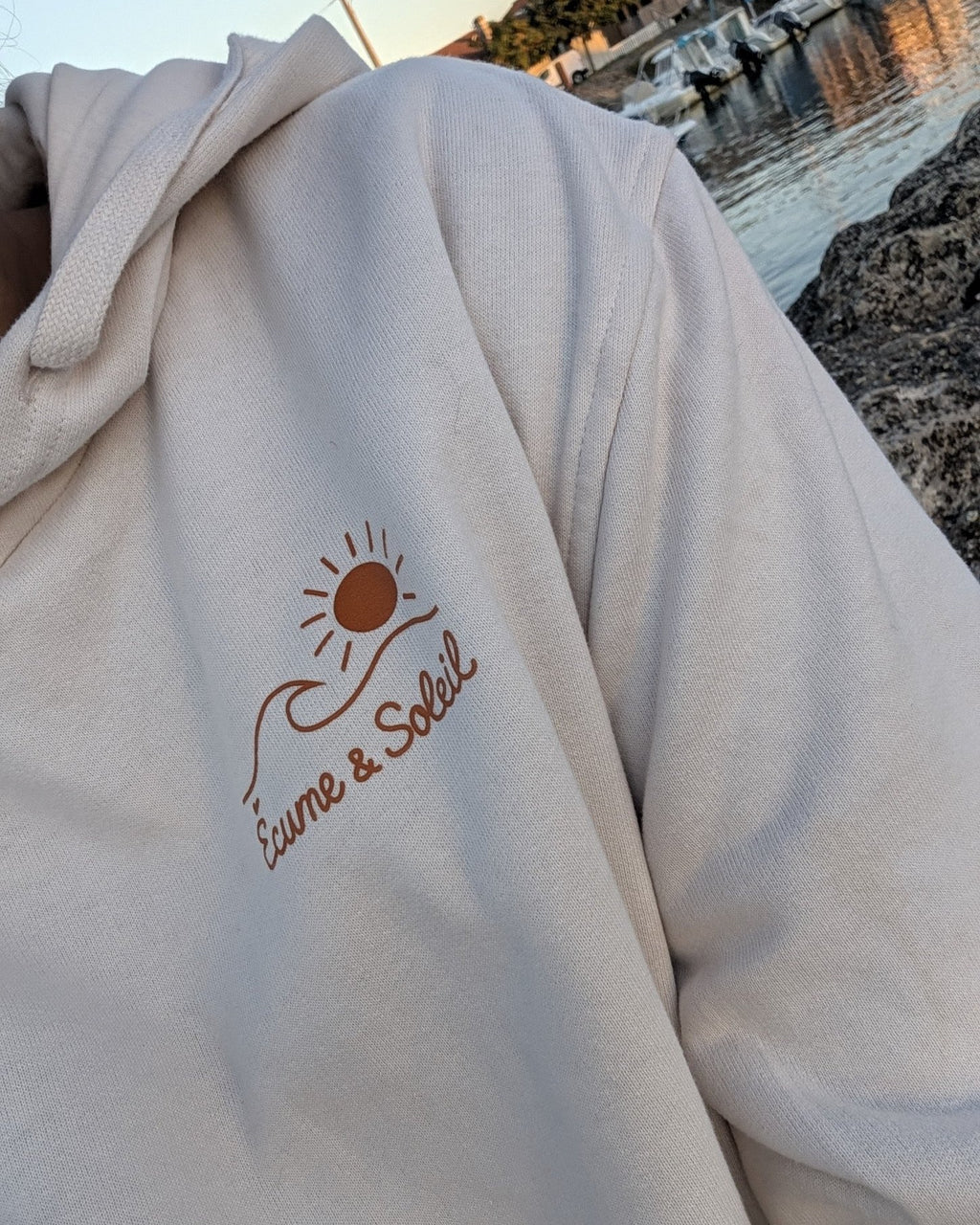 Sweat à capuche Écume & Soleil écru porté au port, logo soleil et vague et signature marqués au cœur