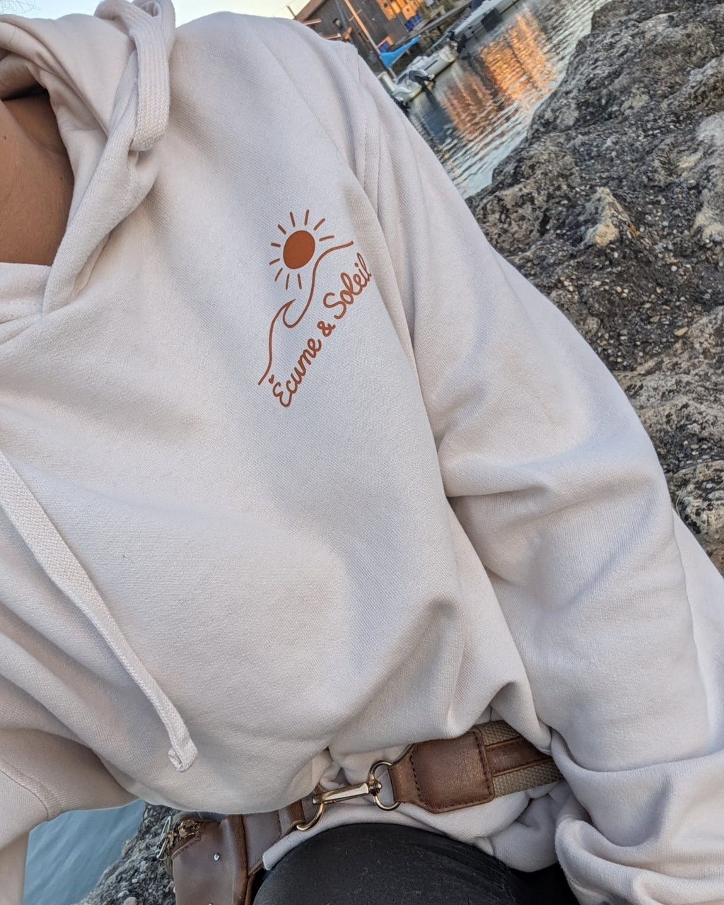 Sweat à capuche écru porté au bord de l'eau, logo Écume & Soleil marqué au cœur