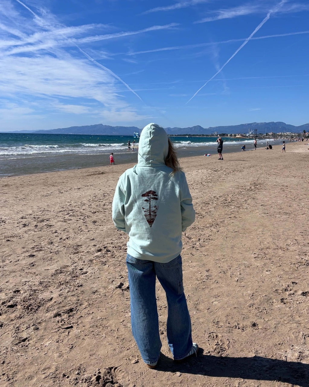 Sweat à capuche Île & Pins blush mint porté de dos face à la mer, avec un jean, motif sépia au centre du dos
