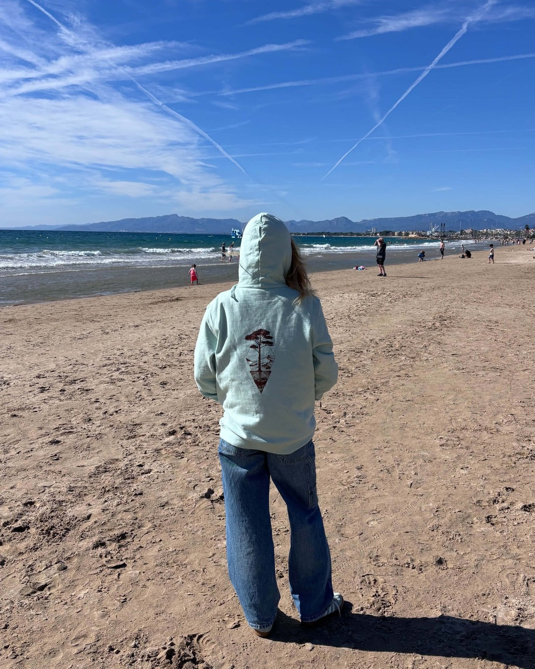 Sweat à capuche Île & Pins blush mint porté de dos face à la mer, avec un jean, motif sépia au centre du dos