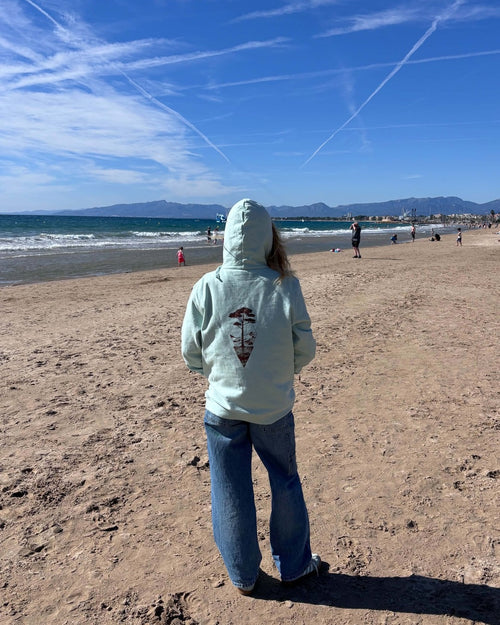 Sweat à capuche Île & Pins blush mint porté de dos face à la mer, avec un jean, motif sépia au centre du dos