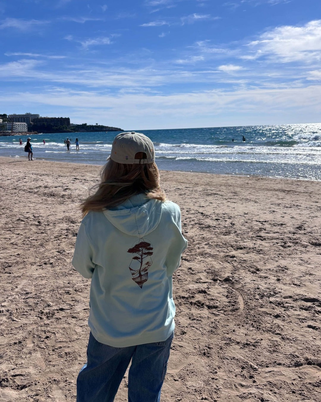 Sweat à capuche Île & Pins blush mint porté de dos sur la plage, motif pin et pinasse en sépia