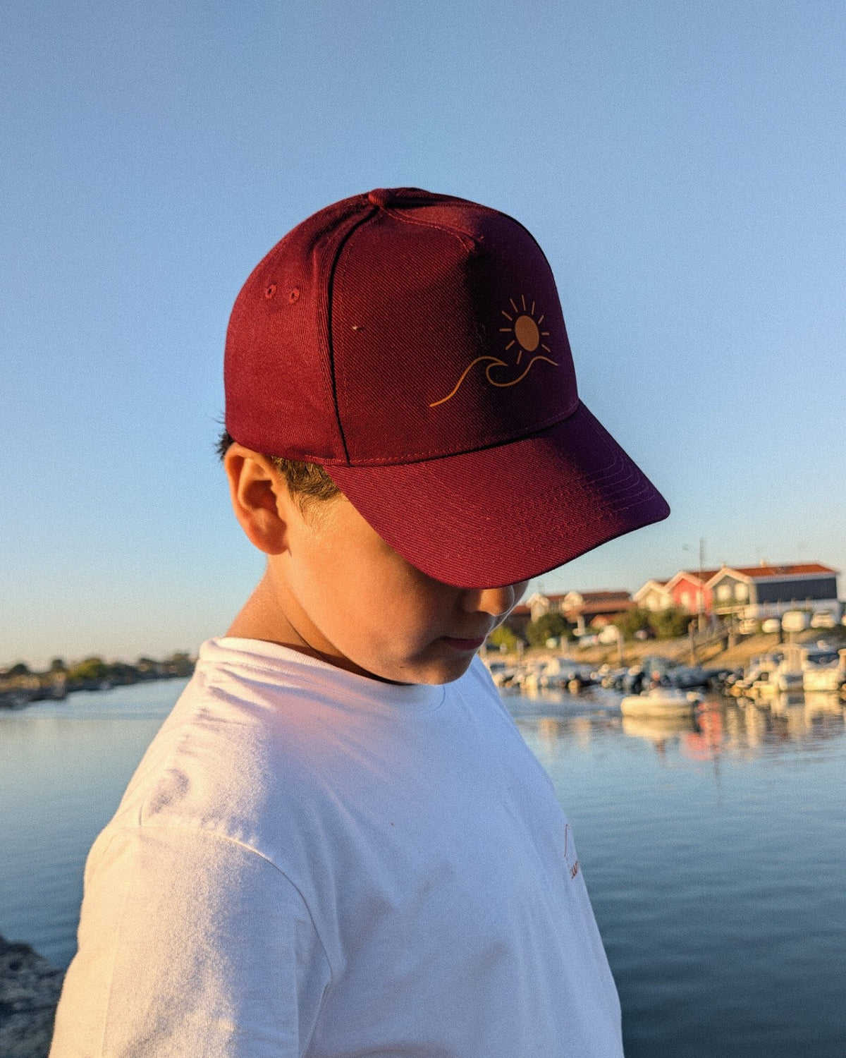 Casquette Écume & Soleil bordeaux portée au port, logo soleil et vague marqué en orange