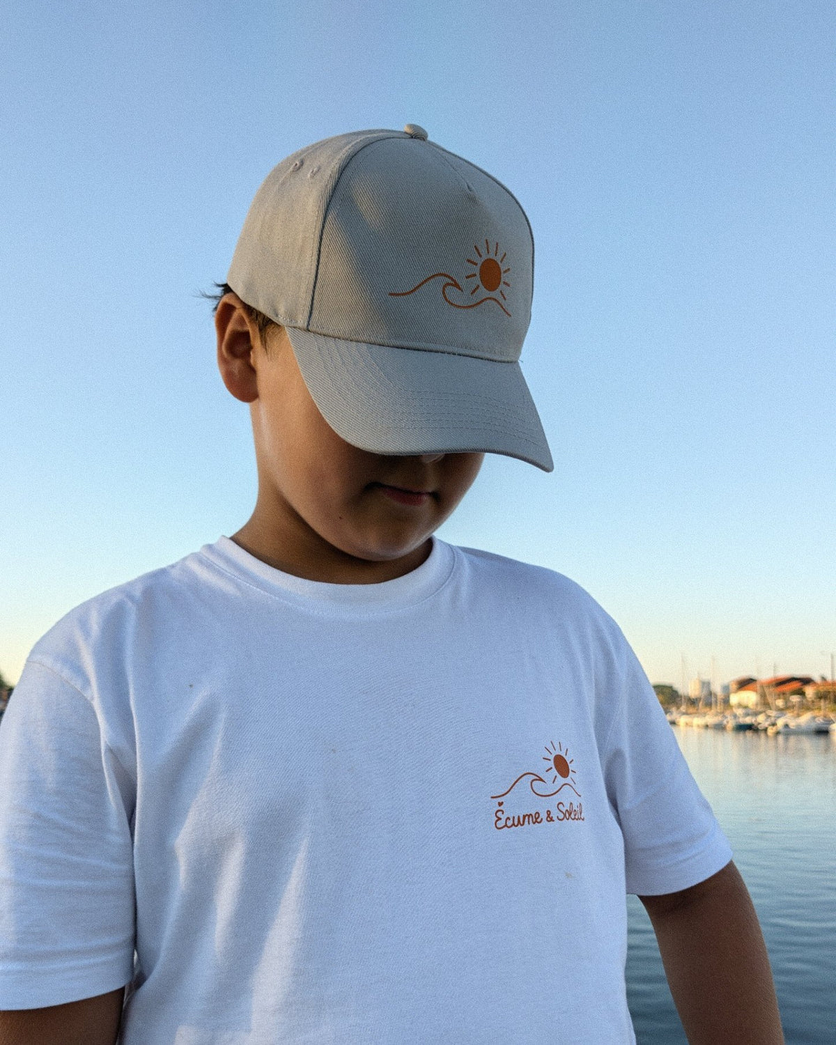 Casquette Écume & Soleil gris clair portée au port, logo soleil et vague en orange