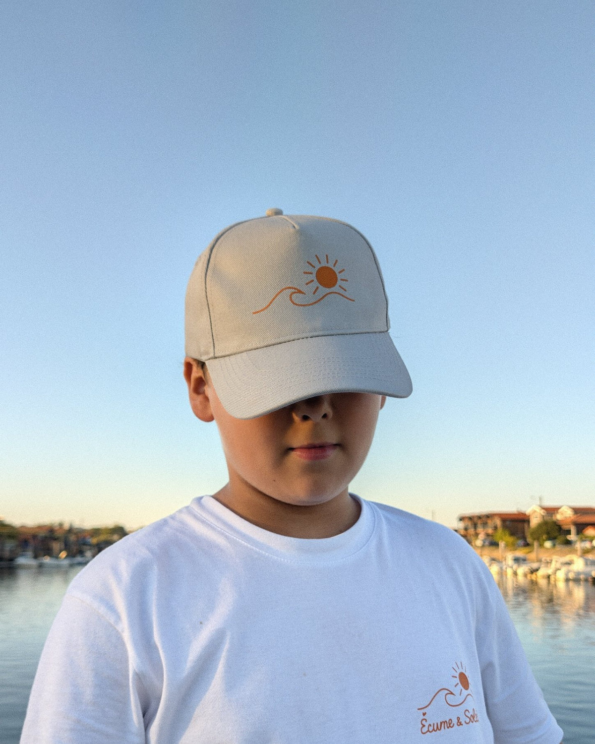Casquette Écume & Soleil gris clair vue de face au bord de l'eau, logo soleil et vague