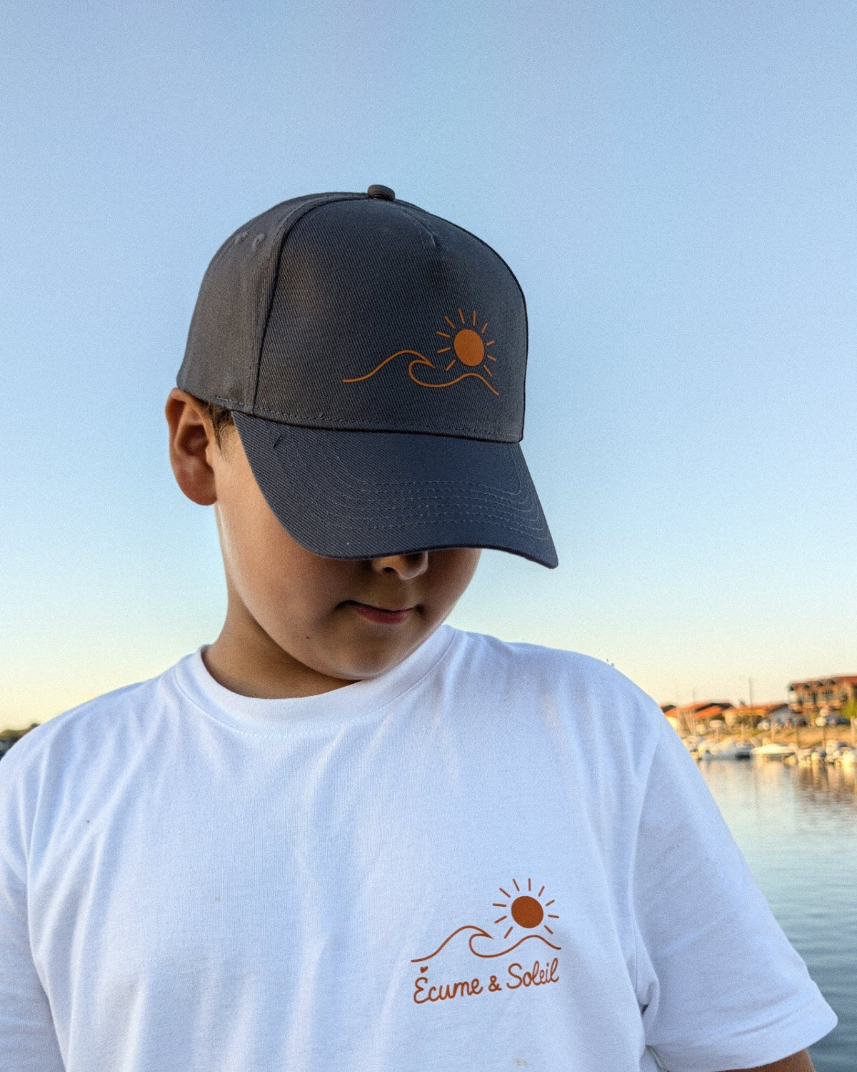 Casquette Écume & Soleil gris foncé portée au port, logo soleil et vague en orange