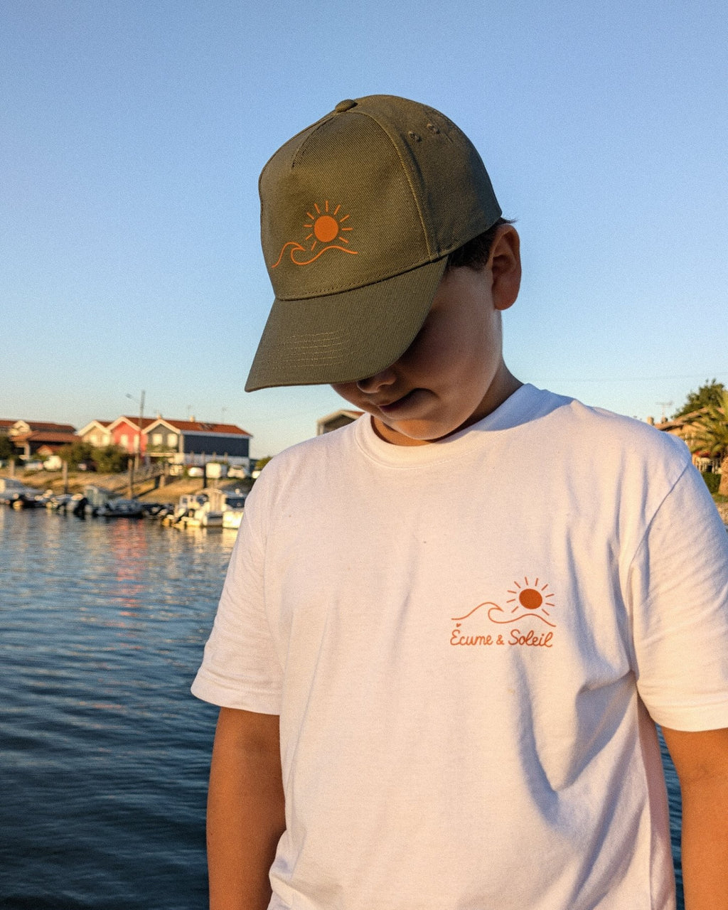 Casquette Écume & Soleil kaki portée au port, logo soleil et vague marqué en orange, avec le t-shirt blanc logo au cœur