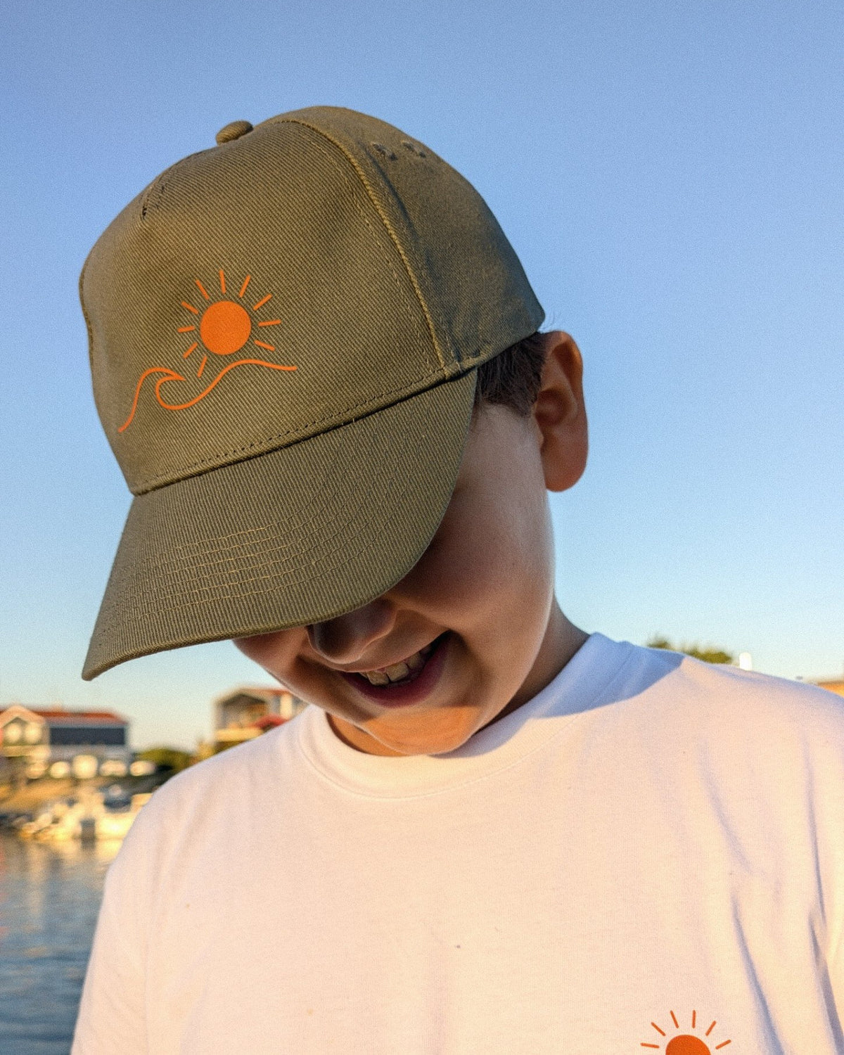 Casquette Écume & Soleil kaki, gros plan du logo soleil et vague, au coucher du soleil sur le port