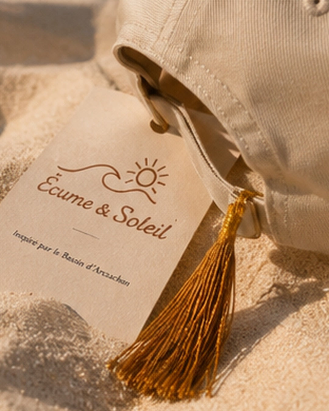 Étiquette Écume & Soleil « Inspiré par le Bassin d'Arcachon » et pompon doré posés sur le sable avec la casquette écrue