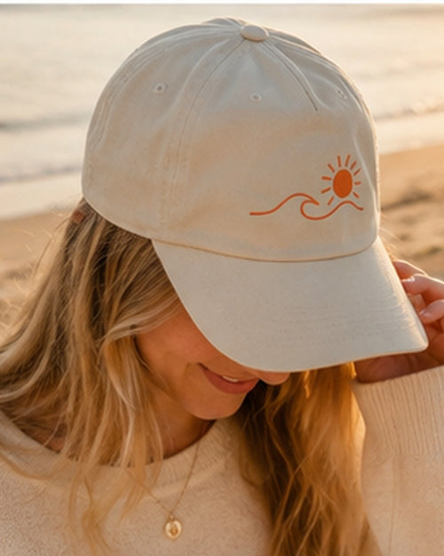 Casquette Écume & Soleil écrue portée sur la plage au coucher du soleil, logo soleil et vague orange