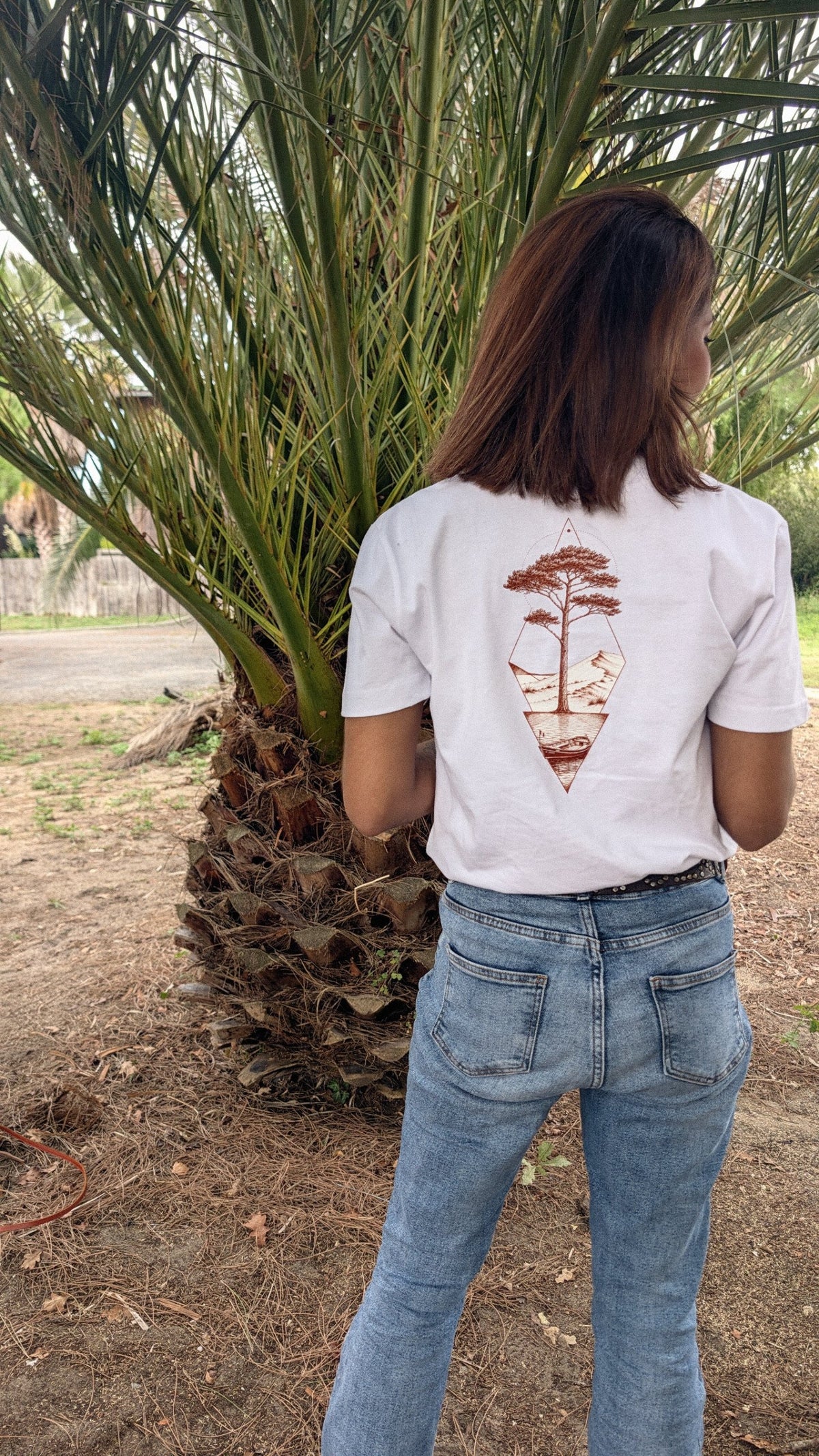 Femme portant le T-shirt Écume & Soleil Île & Pins blanc, vue de dos : le pin, la dune et la pinasse marqués en sépia au centre du dos, sous un palmier