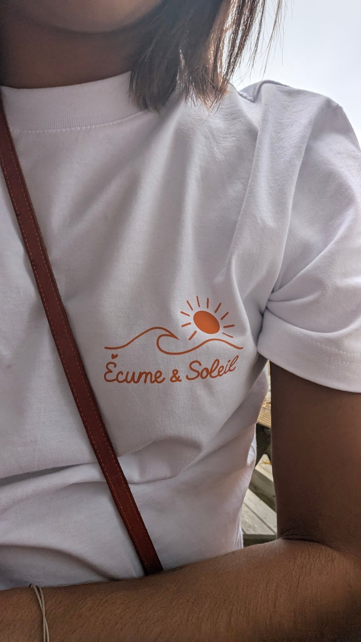T-shirt Écume & Soleil Île & Pins blanc porté, gros plan du logo soleil & vague et de la signature Écume & Soleil marqués en orange au cœur