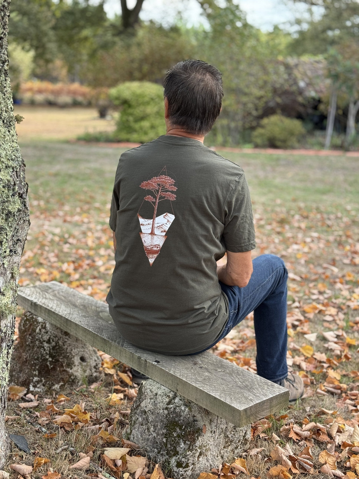 Homme assis sur un banc de jardin, vu de dos, portant le T-shirt Écume & Soleil Île & Pins kaki : le pin, la dune et la pinasse marqués au centre du dos