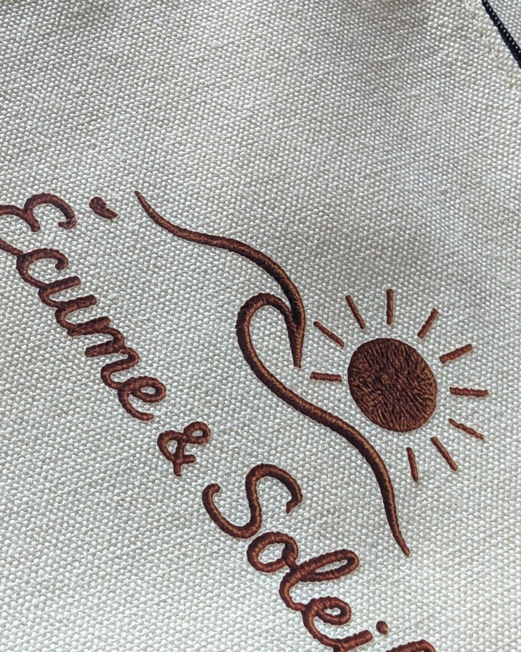 Détail de la signature effet relief sur la pochette écrue : soleil, vague et nom Écume & Soleil en terracotta
