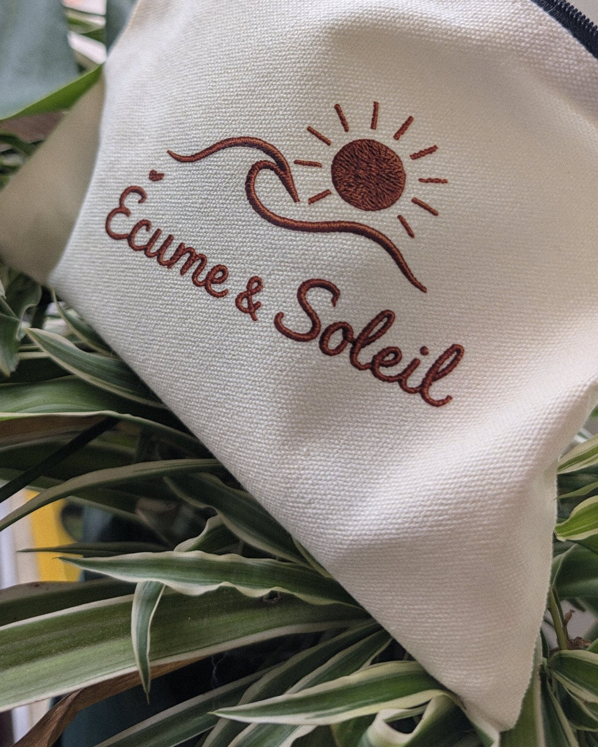 Pochette écrue Écume & Soleil au milieu du feuillage, logo soleil et vague en terracotta