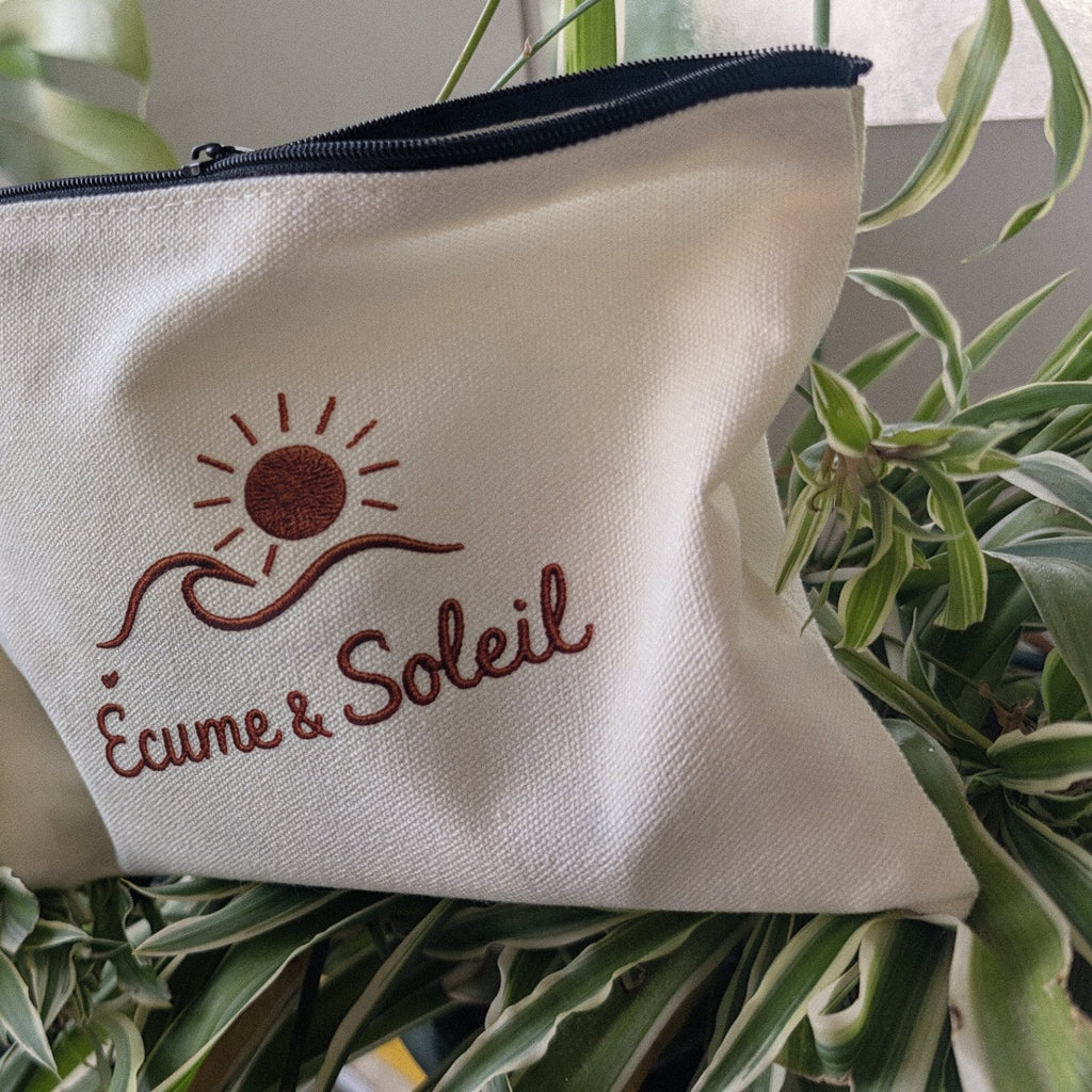 Pochette Libre Ensemble écrue Écume & Soleil posée dans les plantes, zip noir, signature soleil, vague et nom en terracotta effet relief
