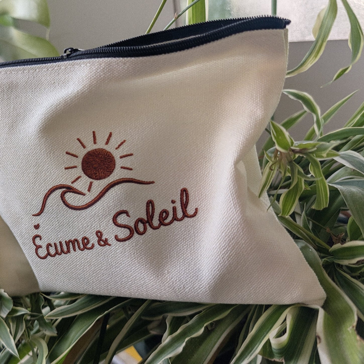 Pochette Libre Ensemble écrue Écume & Soleil posée dans les plantes, zip noir, signature soleil, vague et nom en terracotta effet relief