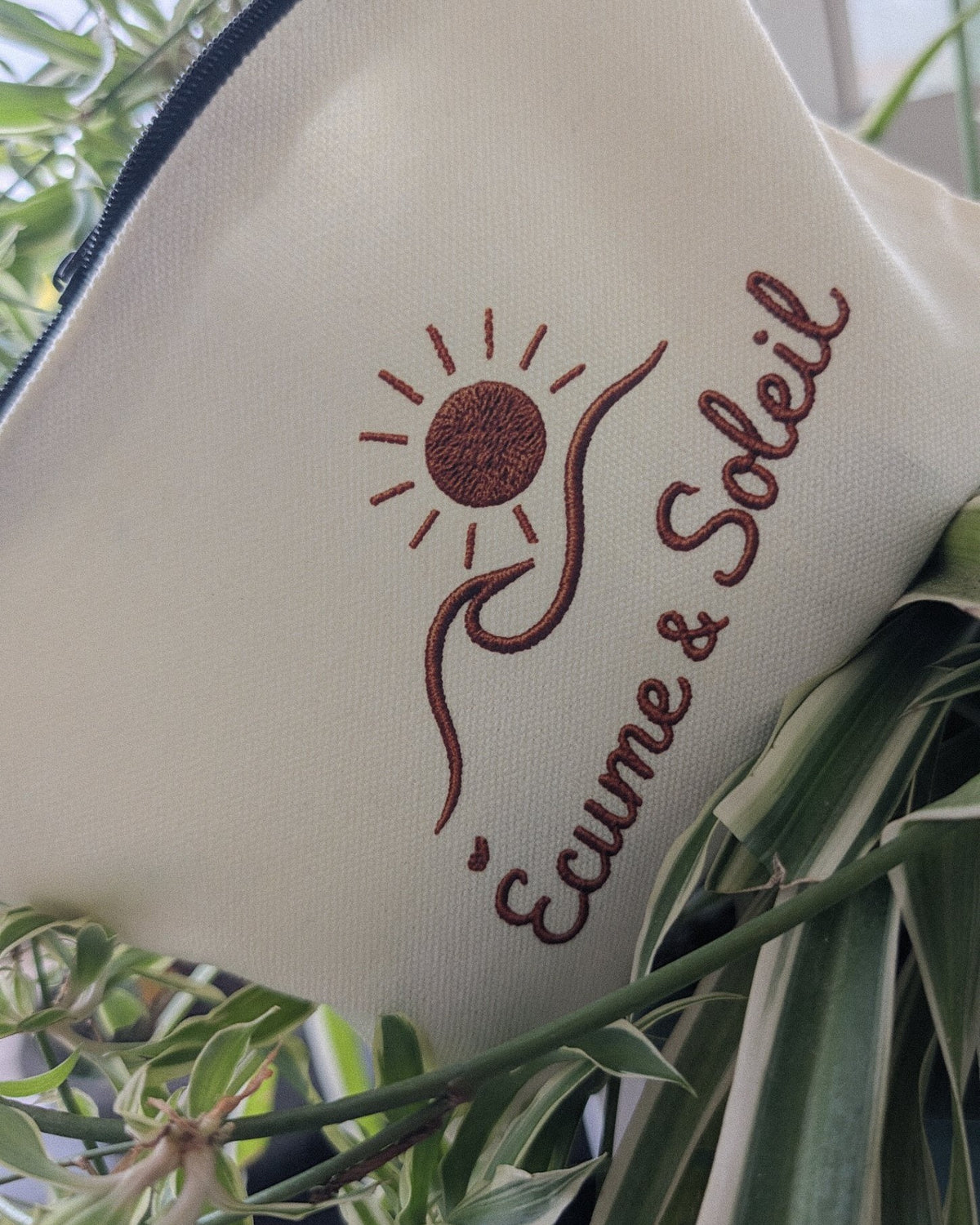 Gros plan de la signature effet relief Écume & Soleil sur la toile écrue de la pochette : soleil, vague et nom en terracotta