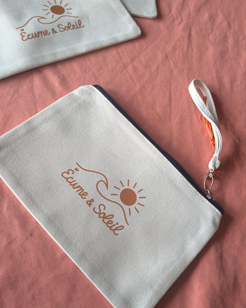 Pochettes Écume & Soleil écrues sur une nappe, zip marine, signature soleil, vague et nom en terracotta