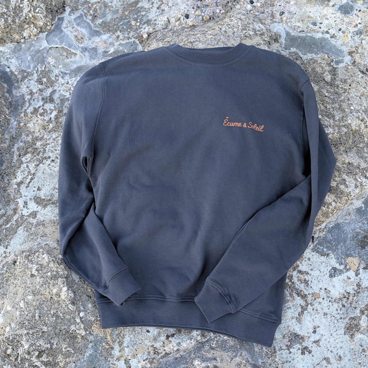 Pull Le Basique Écume & Soleil anthracite à plat, nom marqué au cœur, sur les rochers du Bassin d'Arcachon