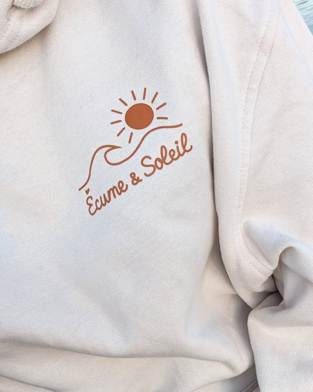 Gros plan du logo Écume & Soleil effet relief au cœur du sweat à capuche