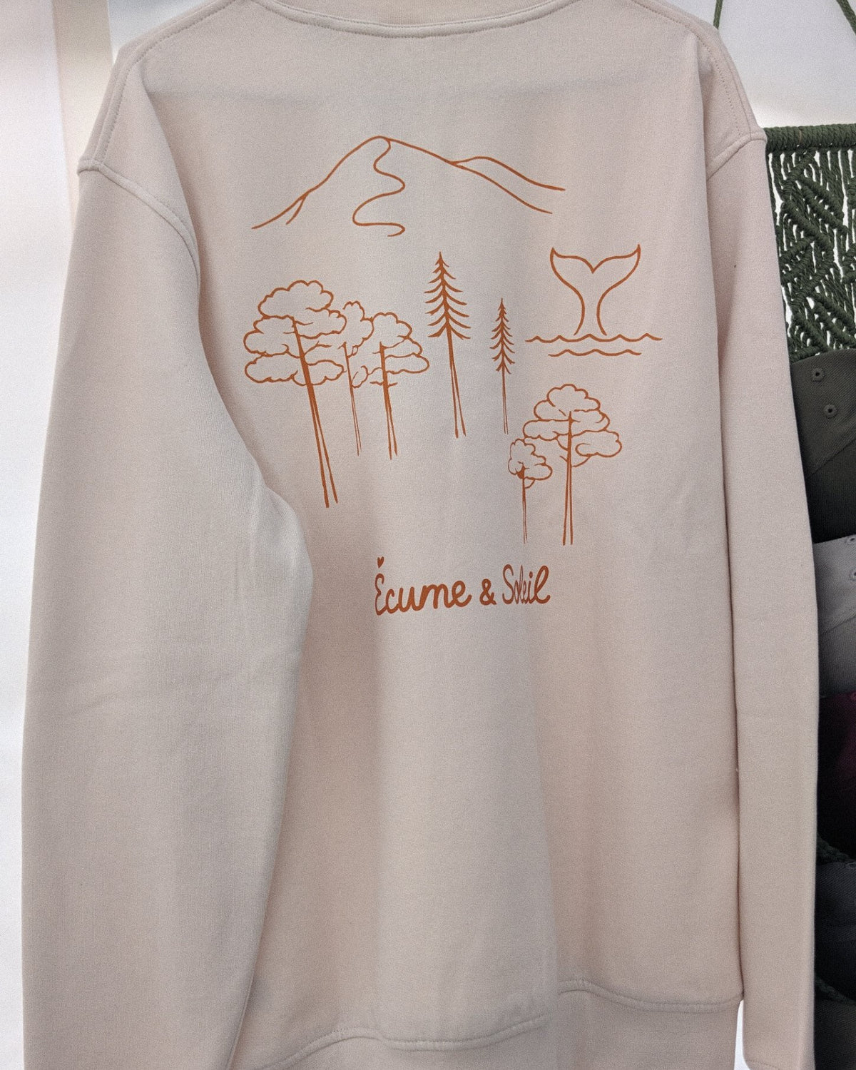 Sweat Écume & Soleil Esprit Bassin écru, vu de dos sur le portant : forêt de pins, dune et queue de baleine, signature sous le motif
